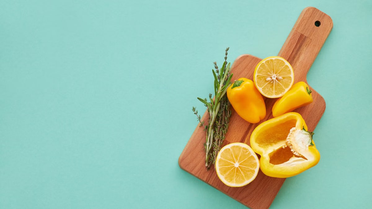 A vibrant display of yellow peppers, lemons, and herbs on a wooden board against a teal background.